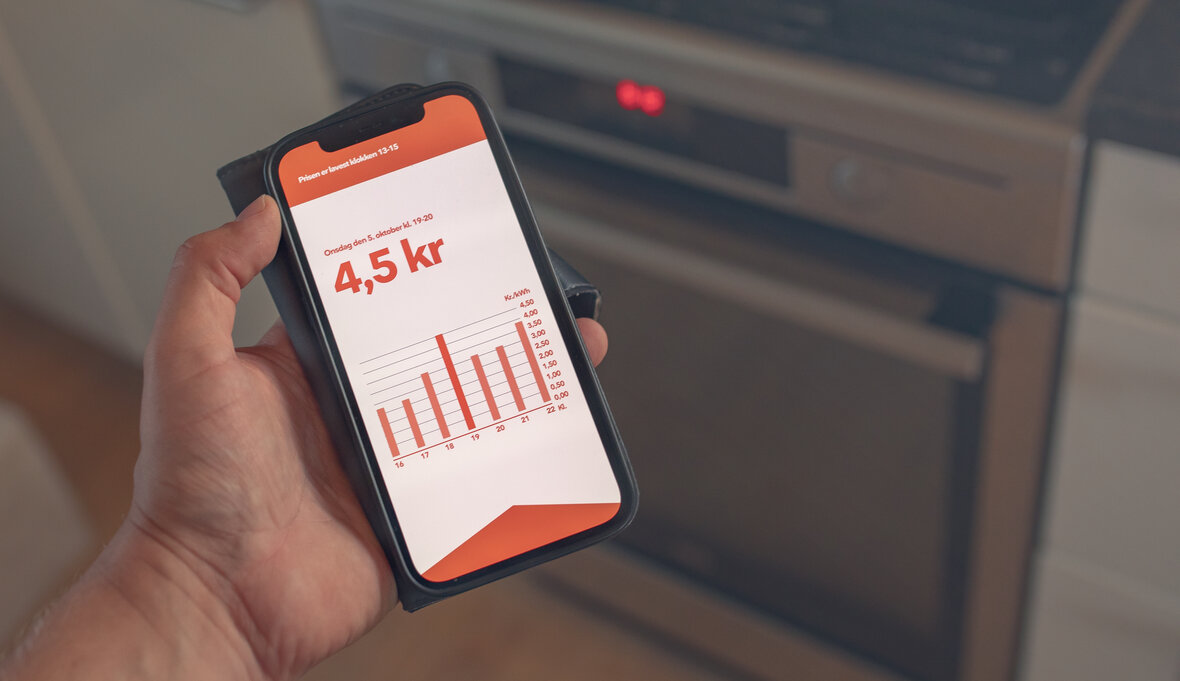 Hånd med mobiltelefon foran en ovn. På mobilens skærm tjekkes elprisen.