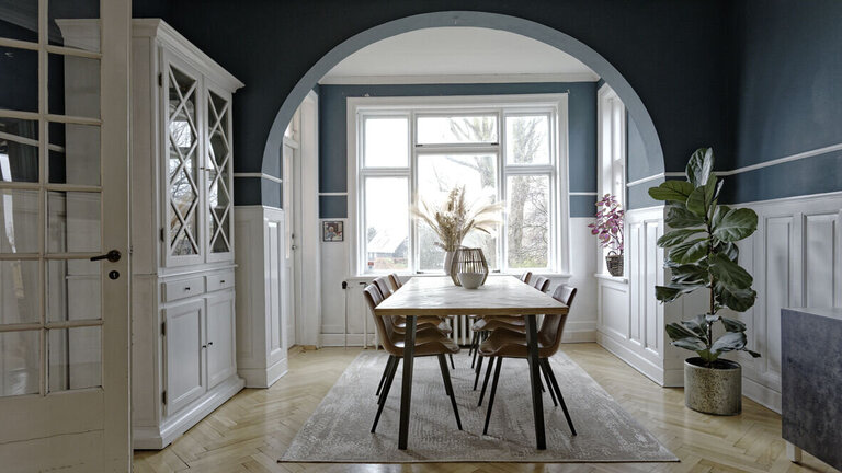 Spisebord ved bue i væggen og stort vinduesparti. Væggene er malet i en petroleumsblå vægfarve