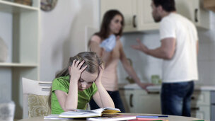 far og mor skændes bagved barn, der sidder ved køkkenbord og holder sig for ørerne