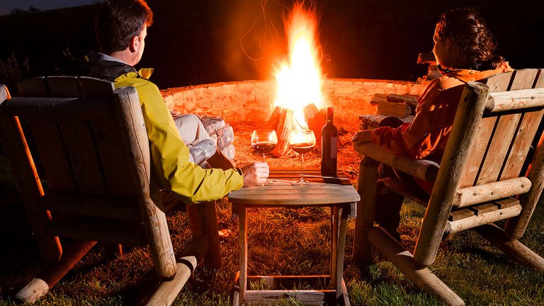 par sidder i haven foran bål og drikker rødvin