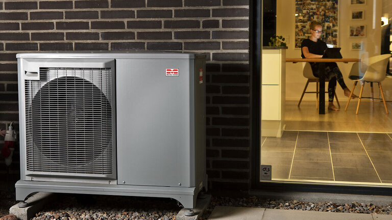 Varmepumpe foran hus, hvor kvinde sidder ved spisebord
