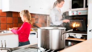 Metalgryde på kogeplade med kvinde og pige i baggrunden.