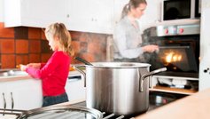 Metalgryde på kogeplade med kvinde og pige i baggrunden.